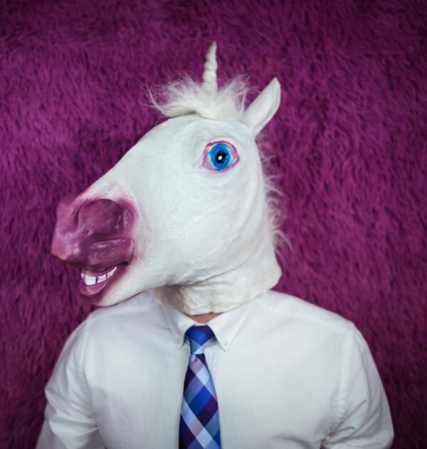 Freaky young man in comical mask stands on the purple background. Portrait of unusual manager. Serious unicorn in shirt and tie is looking away