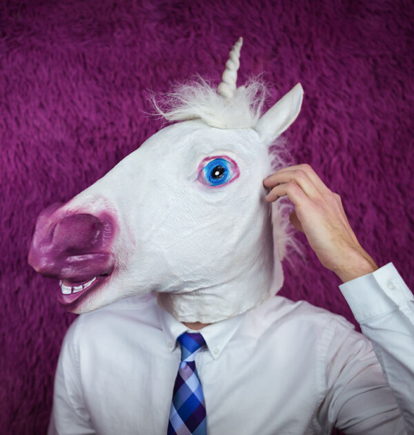 Freaky young man in comical mask stands on the purple background. Portrait of unusual manager. Confused unicorn in shirt and tie is thinking